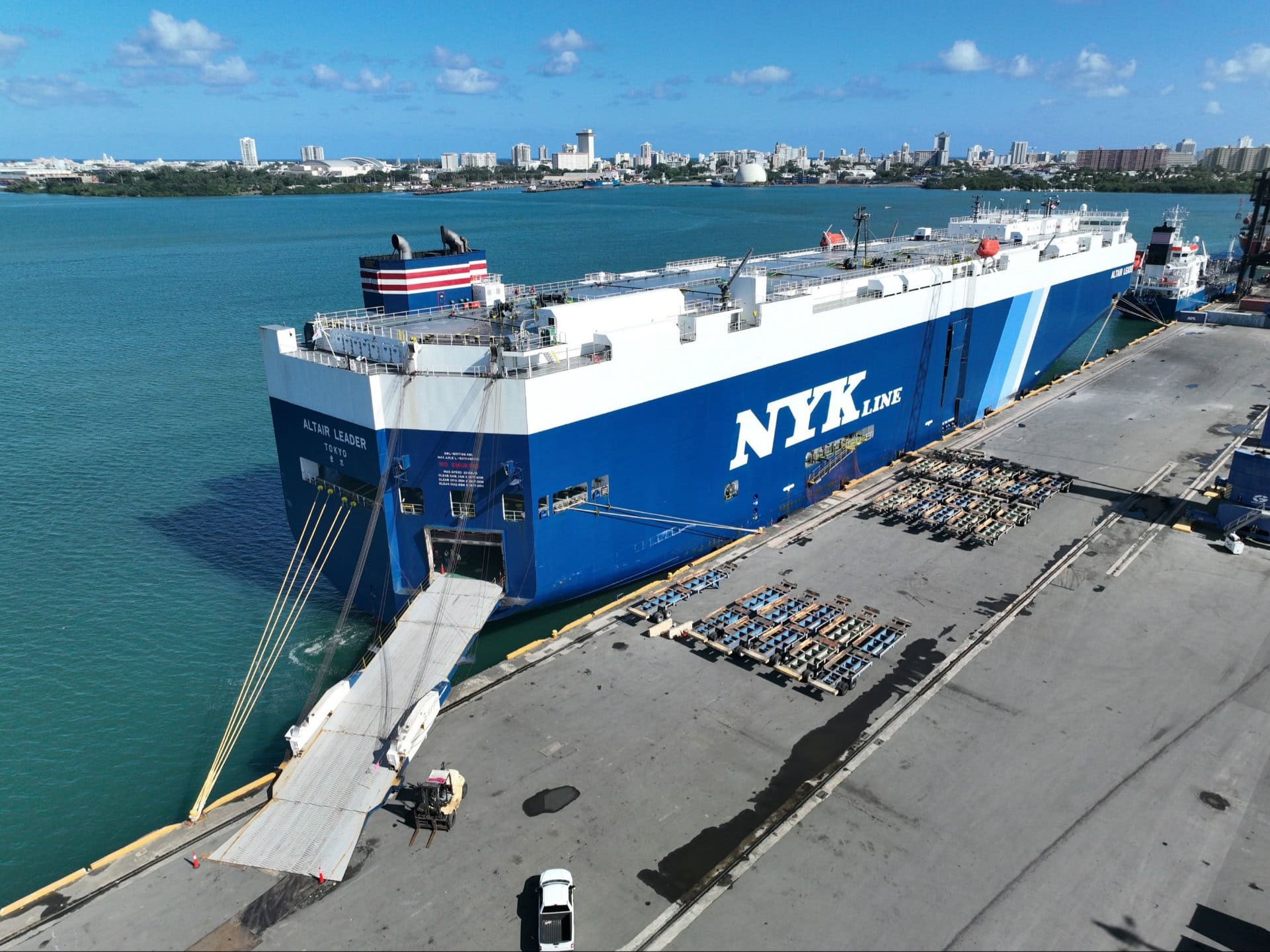 NYK Line cargo ship docked at Puerto Rico terminal with port infrastructure and city skyline in back.