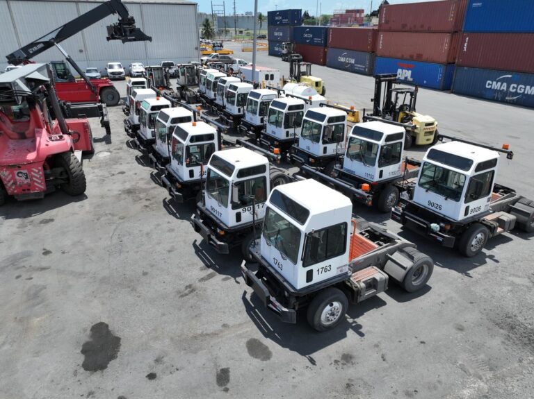 Small fleet of white utility vehicles parked in a lot with shipping containers in the background.